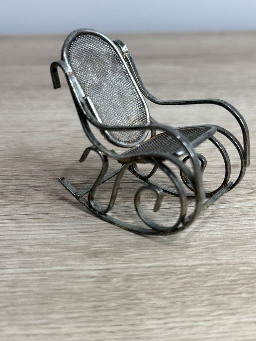 A small, metal rocking chair with a mesh backrest sits on a wooden surface.