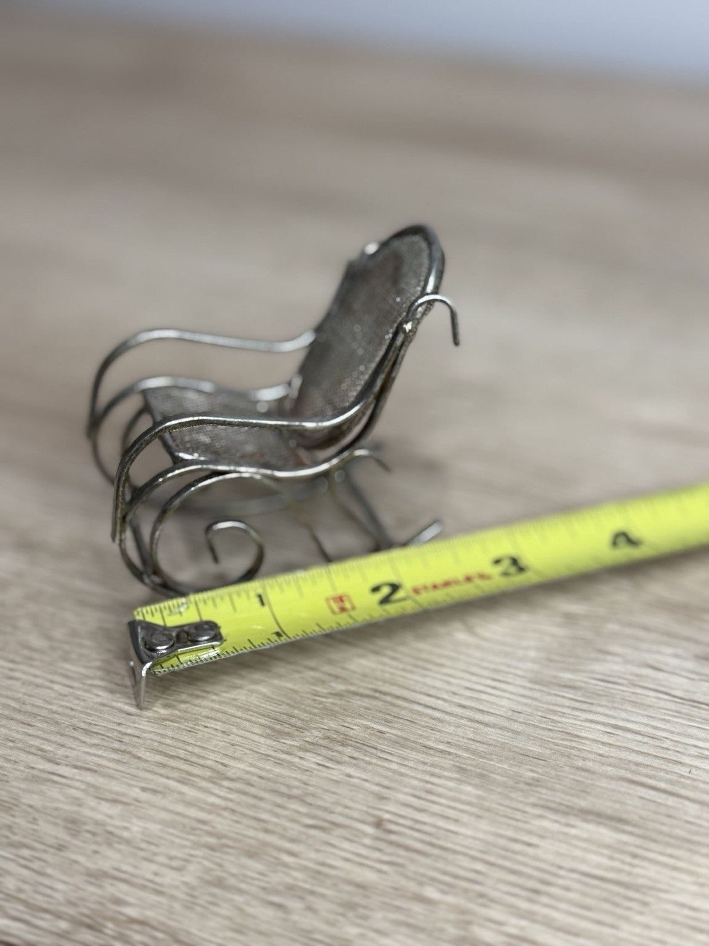 A small metal rocking chair is placed next to a yellow measuring tape on a wooden surface.