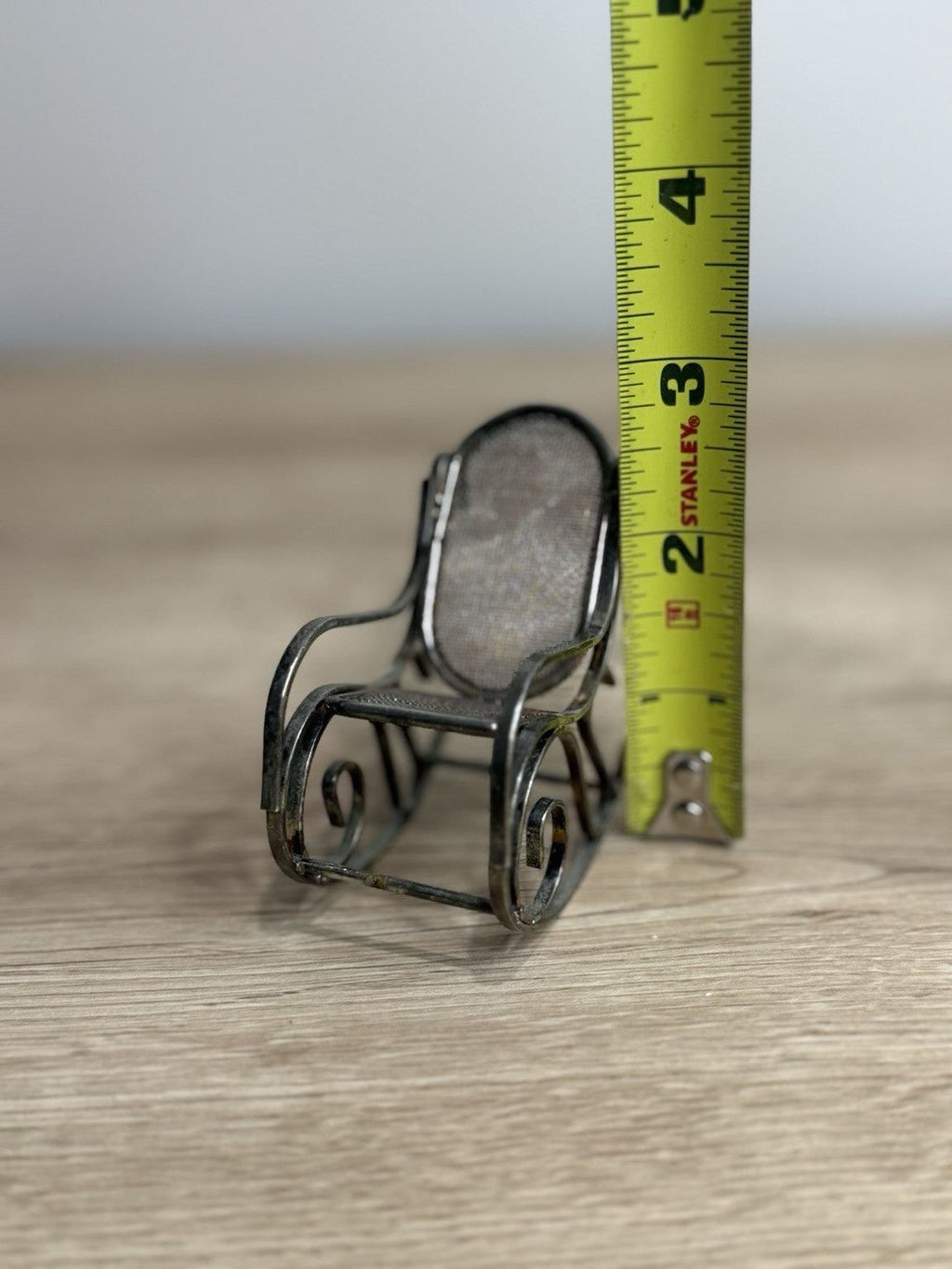 A small metal rocking chair is placed next to a yellow measuring tape on a wooden surface.