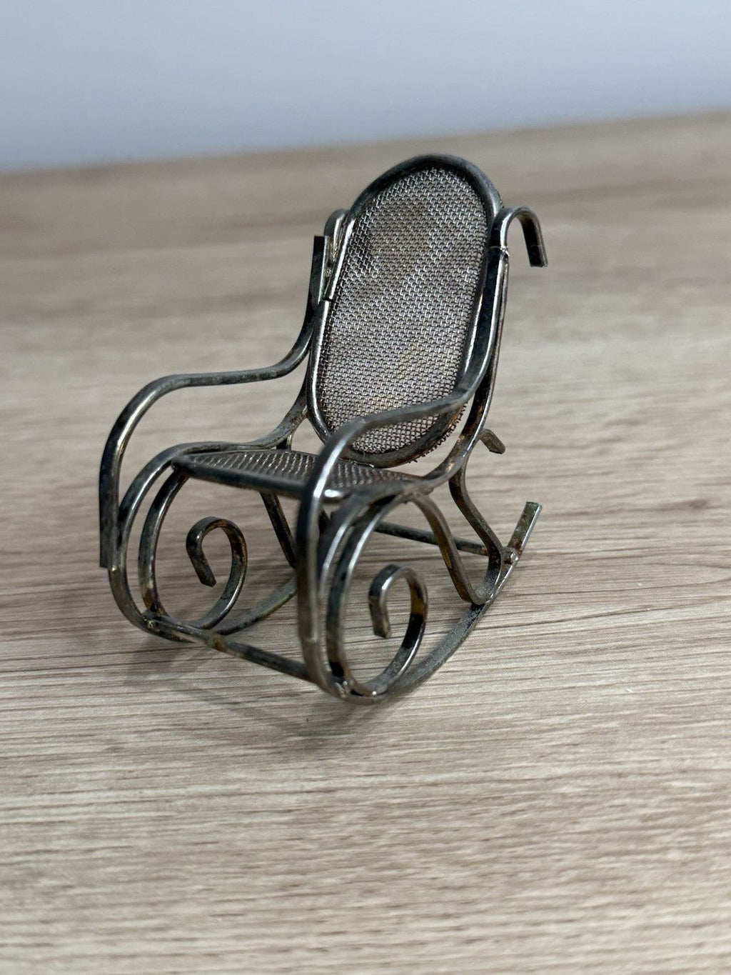 A small, metal rocking chair with a mesh backrest sits on a wooden surface.